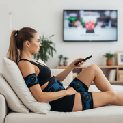 Woman sitting on a couch wearing black and blue massage wraps byGYM IN A BOX holding a remote control with a TV in the background.