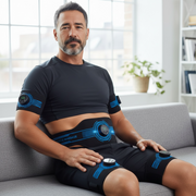 Man wearing a black and blue GYM IN A BOX EMS massage device on a couch in a bright room.