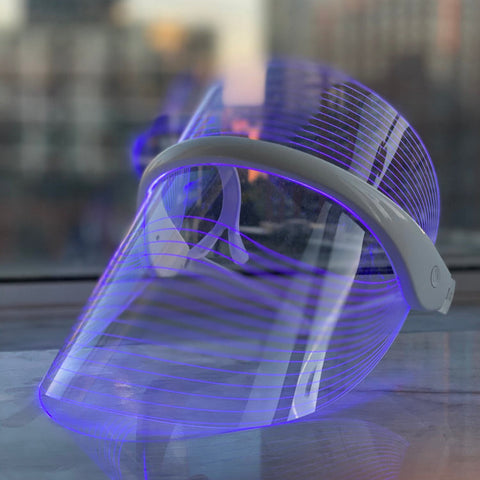 Magic Glow LED BeautyShield mask glowing with blue light therapy, designed to target acne and inflammation; displayed on window ledge with cityscape view.
